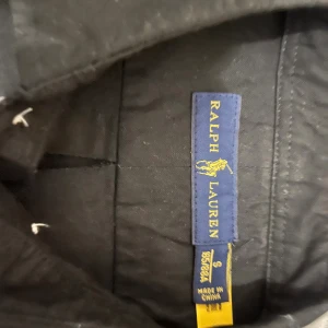 Svart skjorta från Ralph Lauren - Svart skjorta från Ralph Lauren med klassisk krage och vita knappar framtill. På bröstet finns den ikoniska röda Polo-loggan broderad. Skjortan är gjord i mjuk bomull och har lång ärm, perfekt för en clean och stilren look.