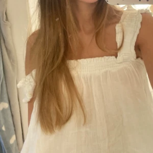 Vit somrig blus med breda axelband - Supersöt vit blus med breda axelband och överdel. Blusen har en luftig, flowy passform som känns perfekt för varma dagar. Materialet är lätt och mjukt, troligen bomull eller linne, vilket gör den extra skön att bära.