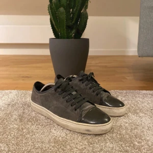 Lanvin - Snygga gråa sneakers från lanvin med ovandel i mocka och blanka detaljer på tå och häl. Skorna har svarta snören och en vit, platt sula som ger en clean look. Perfekta för dig som gillar en stilren och urban vibe. Skick 8/10. Skriv vid frågor eller prisförslag!😁