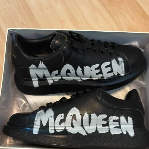 Alexander McQueen svarta sneakers - Svarta sneakers från Alexander McQueen i skinn med fet vit McQueen-grafitti på sidan. Klassisk chunky sula, svarta skosnören och diskreta gulddetaljer på plösen. Riktigt statement-sneakers för dig som vill sticka ut.