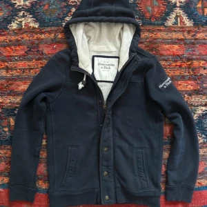 Svart hoodiejacka Abercrombie & Fitch - Snygg svart hoodiejacka från Abercrombie & Fitch med dragkedja och knappar framtill. Jackan har huva med vita snören, två fickor och ett broderat märke på ärmen. Insidan är mjukt fodrad i beige och materialet är bomull. Perfekt för chill och streetwear.