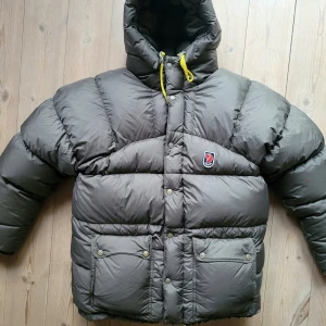 XS Unisex - Grå dunjacka från Fjällräven med huva - Säljer en grå dunjacka från Fjällräven med gul snörning i huvan och snygga tryckknappar framtill. .Jackan är tvättad fackmannamässigt, så är ren, fräsch och fluffig. De skador/skavanker som ska nämnas är; - ytterkant på nudlarna på ärmarna är lite slitna, se sista bilden. Kan lagas hos skräddare (prisuppgift till mig 300-500 kr).  - läderflärparna på gula dragbanden är slitna, men man kan få nya vita gratis via Fjällrävens kundtjänst