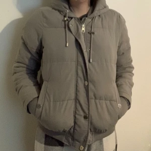Grå pufferjacka - Grå pufferjacka med huva. Jackan har ribbade muddar och två fickor framtill. Perfekt till vintern då den är varmfodrad. Snörena finns inte på jackan längre