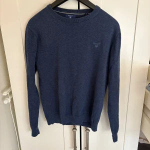Marinblå tröja från GANT - Snygg marinblå stickad tröja från GANT i storlek M. Tröjan har rund hals, diskret logga broderad på bröstet och långa ärmar. Perfekt för dig som gillar klassisk och stilren design med en mjuk känsla.