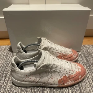 Maison Margiela vita sneakers med röd färg - Maison Margiela sneakers i vit canvas med röd färgstänk och slitna detaljer. Låg modell med rund tå, snörning och platt sula. Unik design med konstnärlig touch och råa kanter. Snygga att styla till streetwear eller för att sticka ut i mängden.