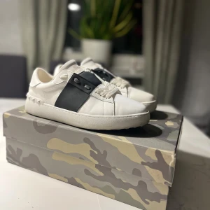 Valentino open storlek 41, passar 42  - Snygga vita Valentino Garavani Rockstud sneakers med bred svart läderdetalj över snörningen och ikoniska nitar vid hälen. Platt sula och rund tå, tillverkade i skinn. Kommer med originalkartong, dustbag och kvitto. Perfekt för dig som gillar exklusiva sneakers med edge.