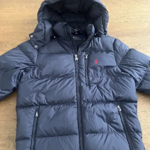 Svart pufferjacka från Polo Ralph Lauren - Snygg svart pufferjacka från Polo Ralph Lauren med huva och flera dragkedjefickor. Jackan har det klassiska röda Polo-logot på bröstet och är tillverkad i polyester. Perfekt för kalla dagar med sin vadderade design och stilrena look.