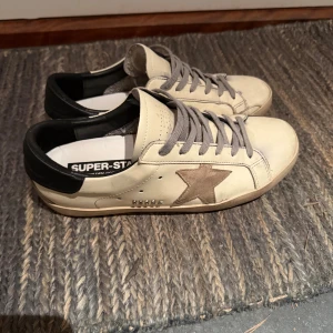 Golden Goose Superstar sneakers beige - Golden Goose Superstar sneakers i beige skinn med grå snörning, svart häl och en ikonisk stjärna i mocka på sidan. Snygg GGDB-detalj i silver och klassisk platt sula. Perfekta för dig som gillar en clean men ändå edgy look. De är bra skick på dom förutom i tån och hälen då dom är lite mer slitna och därför säljer jag också för ett lågt pris