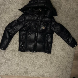 Mörkblå Moncler mont beliard dunjacka med huva - Mörkblå dunjacka från Moncler Mont Beliard med glansig finish, huva och klassisk logga på bröstet. Jackan har dragkedja framtill, quiltad design och innerficka med serietidningsmönster. Perfekt för kalla dagar och har en riktigt snygg streetstil.
