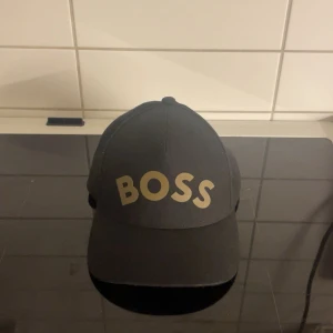 Svart keps med guldtext BOSS - Svart keps från BOSS med justerbart spänne bak och stor guldfärgad text 'BOSS' framtill. Klassisk böjd skärm och stilren design som ger en cool och självsäker look. Perfekt accessoar för att lyfta din outfit.