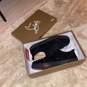 Christian Louboutin svarta sneakers med nitar - Säljer ett par svarta sneakers från Christian Louboutin i mocka med coola svarta nitar på tån och sidorna. Skorna har klassisk röd sula och snörning framtill. Kommer med originalkartong och dustbag. Perfekta för dig som vill sticka ut med lyxig streetstyle. Ingen aning om det är äkta fick det i julklapp och kan inte sådant!