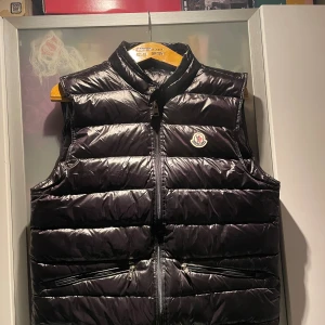 Svart dunväst från Moncler - Snygg svart dunväst från Moncler med glansig finish och klassisk logga på bröstet. Västen har ståkrage, två fickor med dragkedja och quiltad design. Perfekt att slänga över en hoodie för extra värme. Dragkedja framtill för enkel på- och avtagning.
