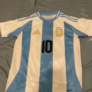 Argentina Messi #10 fotbollströja Adidas - Säljer en officiell Argentina fotbollströja från Adidas med Messi och nummer 10 på ryggen. Tröjan är kortärmad, har klassiska blå och vita ränder, guldiga detaljer och AFA-emblem på bröstet. Tillverkad i lätt och ventilerande Aeroready-material.