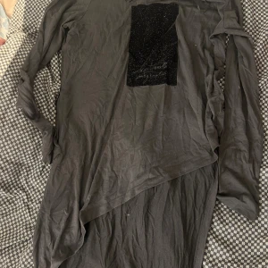 Mörk grå långärmad klänning från Zara - Mörk grå långtopp från Zara med lång ärm och asymmetrisk nederkant. Framsidan har en svart glittrig detalj med text. Tillverkad i mjuk bomull, perfekt för dig som gillar en stilren look med lite edge.