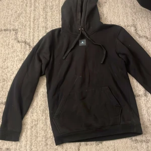 Svart hoodie med elefanttryck - Svart hoodie från Bos Lucas med stor vit patch på ryggen med ett coolt elefantmotiv och texten 'Bos Lucas Et Frigus'. Tröjan har huva med snörning, känguruficka och långa ärmar. Perfekt för dig som gillar unika prints och streetwear.