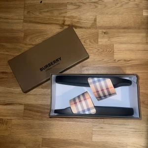 Burberry sandaler med klassiskt rutmönster - Snygga sandaler från Burberry med svart sula och rem i det ikoniska rutmönstret i beige, blått och rött. Perfekta för sommaren och ger en lyxig touch till din outfit. Tillverkade i syntetmaterial med präglad Burberry-logga på sidan och undersidan.