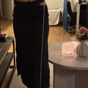 Svart lång kostym kjol med slits och ränder - Säljer en svart lång kostym kjol med tunna i randigt mönster. Kjolen har hög midja, markerade utskurna detaljer vid midjan och en snygg slits på sidan. Passformen är rak.