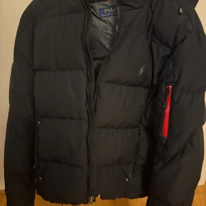 Svart pufferjacka Polo Ralph Lauren M - Svart pufferjacka från Polo Ralph Lauren med broderad logga på bröstet och röd dragkedjedetalj på ärmen. Jackan har huva och klassisk quiltad design, perfekt för kalla dagar. Snygg och sportig look med normal passform.