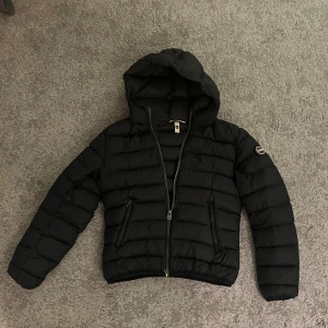 Mörkblå dunjacka med huva från Colmar - Svart dunjacka från Colmar med huva och tvåvägsdragkedja. Jackan har quiltad design, två fickor med dragkedja och Colmar-logga på ärmen. Perfekt för kyliga dagar och riktigt snygg till streetwear-stil.