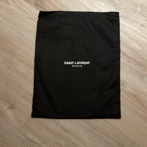 Svart dustbag från Saint Laurent - Svart dustbag från Saint Laurent Paris, perfekt för att förvara din väska eller accessoarer. Dustbagen är rektangulär med vit logotyp framtill och har en enkel snörning upptill. Stilren och klassisk design som passar till alla Saint Laurent-väskor.