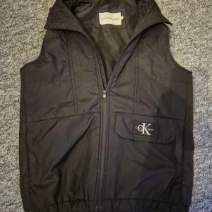 Svart dunväst från Calvin Klein Jeans - Svart dunväst från Calvin Klein Jeans med dragkedja framtill och en ficka med CK-logga på bröstet. Västen har hög krage och elastisk nederkant för extra komfort. Perfekt för lager-på-lager och streetstyle.
