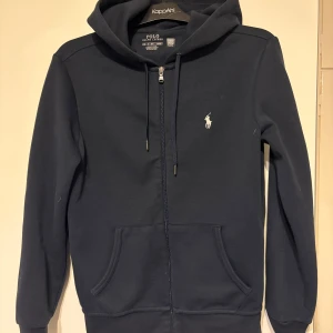 (Xs)Marinblå hoodie från Polo Ralph Lauren - Säljer en marinblå hoodie från Polo Ralph Lauren i storlek XS. Tröjan har dragkedja, huva med snörning och klassisk vit logga på bröstet. Känguruficka framtill och ribbade muddar. Perfekt för en chill och stilren look.