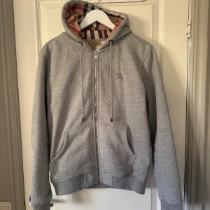 Grå Burberry hoodie med dragkedja - Grå hoodie från Burberry med klassiskt rutigt foder i huvan och insidan. Tröjan har dragkedja, snörning vid huvan och två fickor framtill. Broderad Burberry-logga på bröstet.