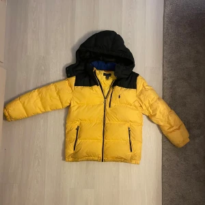 Gul pufferjacka från Polo Ralph Lauren - Grym gul pufferjacka från Polo Ralph Lauren med svarta detaljer och huva. Jackan har dragkedja framtill, två fickor med dragkedja och ikoniska Polo-loggan broderad på bröstet. Perfekt för kalla dagar och riktigt snygg streetstil. L 14-16 passar mellan 150-164 cm
