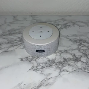 Defunc Bluetooth Speaker - Stilren vit Defunc Bluetooth-högtalare med tydliga knappar för volym och uppspelning. Utrustad med micro-USB-laddning och AUX-ingång. Enheten ser ut att vara i mycket gott skick utan synligt slitage eller repor. Perfekt för trådlös musikuppspelning hemma eller på språng.