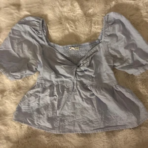 Ljusblå blus med puffärmar och broderi - Supersöt ljusblå blus med puffiga korta ärmar, v-ringning och twistad detalj framtill. Blusen har broderade mönster längst ner och en lätt volangkant. Perfekt för dig som gillar romantisk och trendig stil.