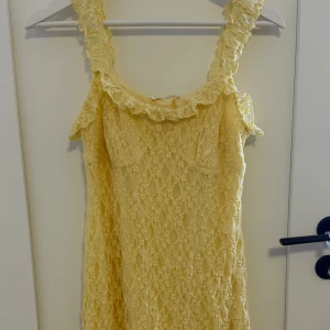 Gul spetsklänning med volangdetaljer - Helt ny supersöt gul klänning med breda axelband och volangdetaljer. Klänningen är täckt av spets och har en romantisk känsla, perfekt för varma dagar. Den är ärmlös och har en kort längd som ger en fräsch och somrig look.