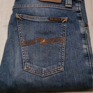 Blå jeans från Nudie Jeans, stl 30/34 - Snygga blå jeans från Nudie Jeans med klassisk femficksdesign och coola orangea sömmar på bakfickan. Jeansen har en rak passform och är tillverkade i slitstarkt denim. Perfekt för dig som gillar stilrena och tidlösa jeans. Slim fit modell