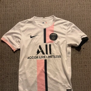 PSG vit och rosa fotbollströja Nike - Säljer en Paris Saint-Germain fotbollströja från Nike i vit med rosa och svart vertikal rand. Tröjan har korta ärmar, rund hals och PSG-logga på bröstet. Materialet är lätt och andas, perfekt för träning eller match. Snygg design med Accor Live Limitless tryck.