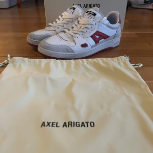 Axel Arigato vita och röda sneakers - Snygga sneakers från Axel Arigato i vitt skinn med röda detaljer och grå mocka-paneler. Klassisk låg modell med snörning och rund tå. Kommer med originalpåse och box. Perfekta för dig som gillar stilrena men ändå unika sneakers.
