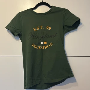 Grön Kingsland Equestrian t-shirt S - Mörkgrön t-shirt från Kingsland Equestrian i bomull med tryck i gult, svart och vitt framtill. Klassisk rund hals och korta ärmar. Perfekt för dig som gillar ridning och vill ha en stilren look med sportig känsla.