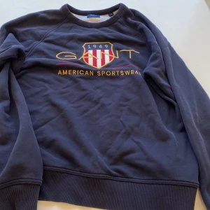 Mörkblå sweatshirt från GANT - Mörkblå sweatshirt från GANT med broderad logga och emblem på bröstet i rött, vitt och gult. Tröjan har rund hals, ribbade muddar och är långärmad. Materialet är mjuk bomull och polyester, perfekt för chill dagar.