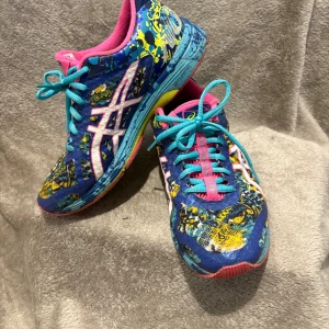 Färgglada Asics sneakers med mönster - Säljer ett par riktigt coola Asics sneakers med blå bas och färgstänk i gult, rosa och turkos. Skorna har snörning, rosa innersula och vit Asics-logga på sidan. Sulan är rosa och blå med detaljer i gult. Perfekta för dig som gillar att sticka ut!
