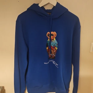 Blå hoodie från Polo Ralph Lauren - Säljer en blå hoodie från Polo Ralph Lauren med ikonisk björn-tryck på bröstet. Tröjan har huva med snörning och långa ärmar. Mjuk bomullskvalitet och klassisk passform. Perfekt för dig som gillar streetwear och snygga detaljer. 