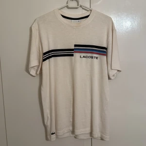 Vit Lacoste t-shirt med ränder - Snygg vit t-shirt från Lacoste i relaxed fit. T-shirten har korta ärmar och ett coolt randigt mönster över bröstet i svart, blått och rött. Materialet är mjuk bomull och loggan syns både på bröstet och vid nederkanten. Perfekt för en chill och stilren look.