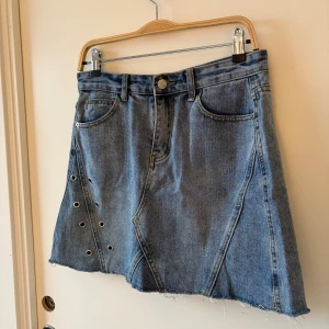 Blå jeanskjol med nitar - Fin blå jeanskjol med rå kant och coola nitar på ena sidan. Kjolen har klassiska fickor fram och bak, bälteshällor och knappstängning. Den har en lätt A-linjeform och är perfekt för dig som gillar en edgy denimstil. Passar en M, liten M