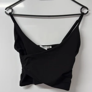 Svart omlott croppad topp NLY Trend - Svart croppad topp från NLY Trend med omlott-design och smala axelband. Toppen är ärmlös och har en snygg v-ringad front. Tillverkad i mjuk bomull med stretch för skön passform. Perfekt att styla med höga jeans eller kjol.