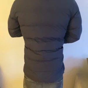 Mörkblå dunjacka från GANT - Säljer en mörkblå dunjacka från GANT med snygg ståkrage och dragkedja framtill. Jackan har quiltad design, två sidofickor och lång ärm. Perfekt för kalla dagar och har en clean look som funkar till jeans och hoodie.