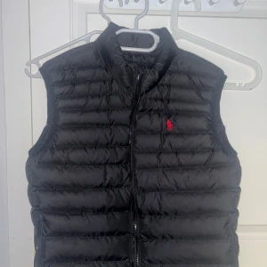 Svart dunväst från Ralph Lauren - Svart quiltad dunväst från Ralph Lauren med klassisk röd logga på bröstet. .