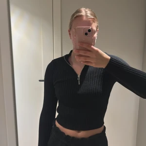 Svart ribbad croppad tröja från Stay - Säljer en svart ribbad croppad tröja från Stay i storlek S. Tröjan har lång ärm, half zip framtill och är i ett stretchigt stickat material som sitter snyggt på kroppen.