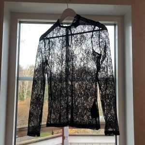 Svart spetsblus från Vero Moda - Säljer en svart långärmad blus från Vero Moda i transparent spets med vackert mönster. Blusen har hög hals med liten knapp i nacken och fina spetsdetaljer runt kragen. Perfekt för dig som gillar en edgy och elegant stil.