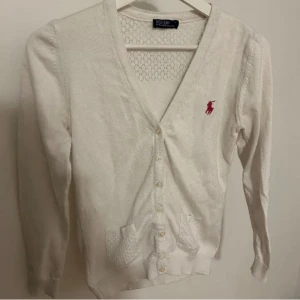 Vit kofta från Ralph Lauren - Vit kofta från Ralph Lauren med v-ringning och knappar framtill. Tröjan har två stickade fickor och den klassiska rosa broderade loggan på bröstet. Långärmad och mjuk i bomull, perfekt för lager på lager under höst och vår. Passar även en person som bär S 