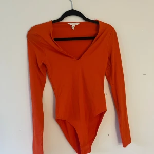 Orange långärmad bodysuit från H&M - Säljer en snygg orange bodysuit från H&M med lång ärm och djup v-ringning. Modellen är tight och har en clean, minimalistisk look som sitter perfekt mot kroppen. Materialet är mjukt och stretchigt, troligen bomull med lite syntet för extra komfort.