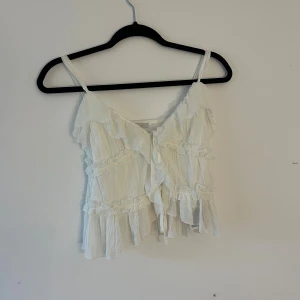 Vit omlott linnetopp med volanger - Supersöt vit linnetopp från Newyorker med tunna axelband, omlott-design och volanger längs hela toppen. Toppen har knyt framtill och är gjord i ett lätt, luftigt material som ger en romantisk vibe. Perfekt för varma dagar och snygg att styla med jeans eller kjol.