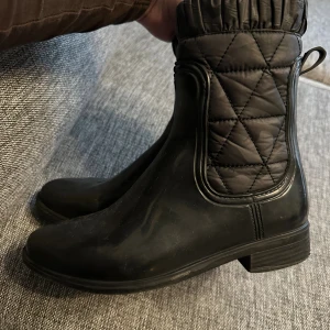 Svarta quiltade boots med platt sula strl: 37 - Säljer ett par svarta boots med quiltad textil upptill och platt sula. Skorna har rund tå, låg klack och är tillverkade i syntetmaterial med vadderad, mönstrad textil på skaftet. Perfekta för höst och vinter när du vill hålla stilen enkel men ändå cool.