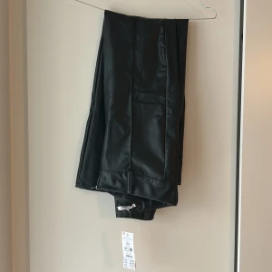 Svarta bootcut PU-byxor från Gina Tricot - Snygga svarta bootcut-byxor i PU-material från Gina Tricot. Byxorna har en cool dragkedja vid benslutet och markerade sömmar framtill. Perfekt för dig som gillar en edgy look och vill sticka ut med stilrena detaljer.